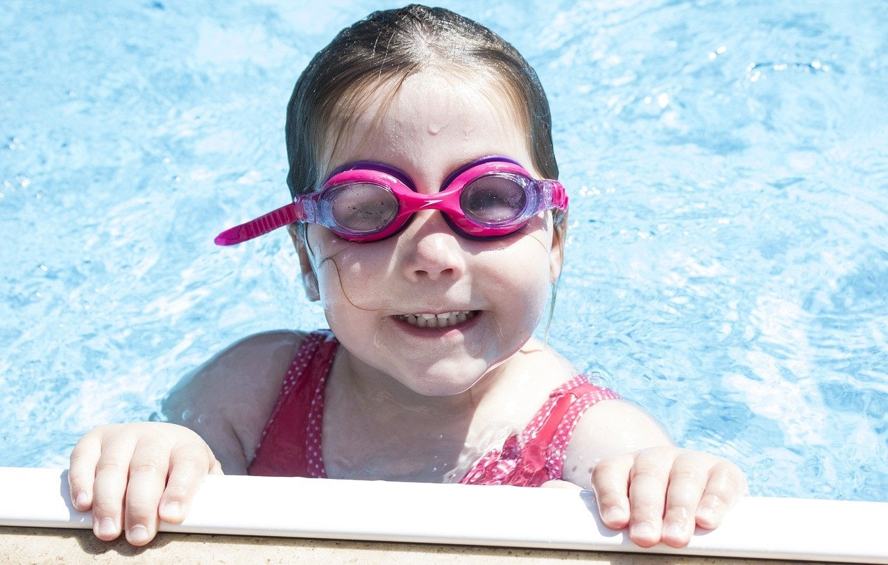 Little girl in goggles hanging onto the side of a pool